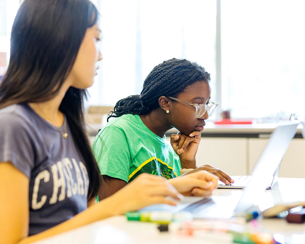 Two students sit at a table, focused on a laptop. One, wearing glasses and a green shirt, looks at the screen intently while the other, in a Chicago t-shirt, types. School supplies are scattered nearby.