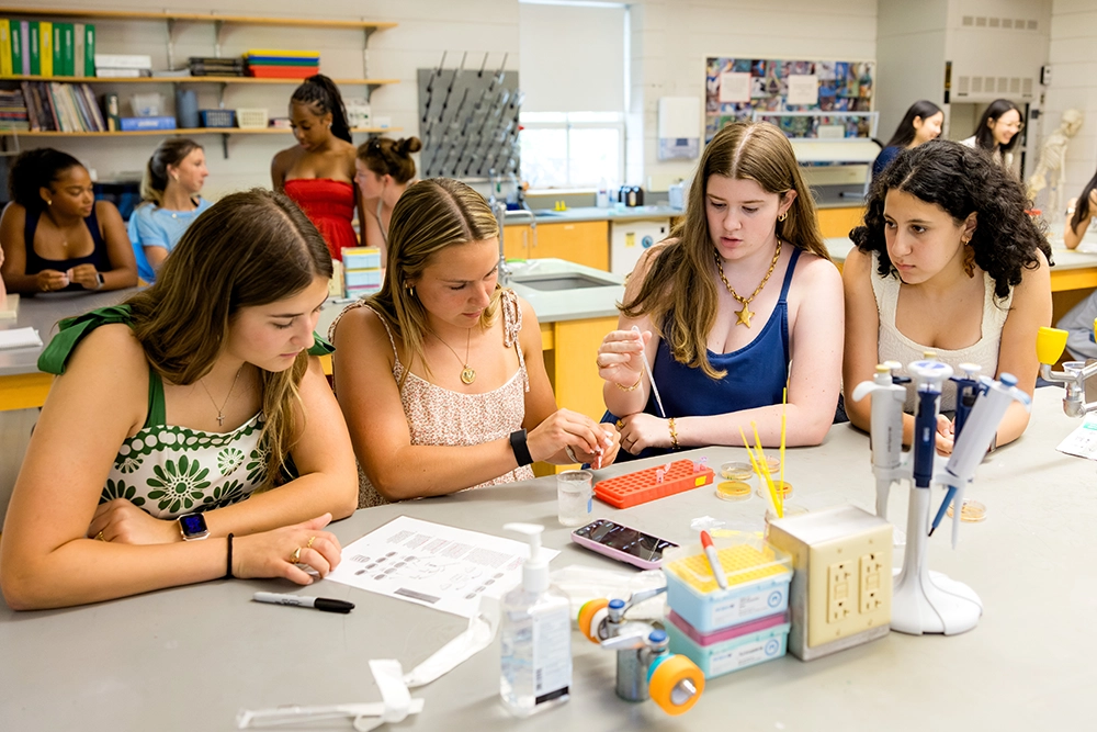 Four young women in a science classroom sit at a lab table, working with pipettes and small containers. Other students and lab equipment are visible in the background.