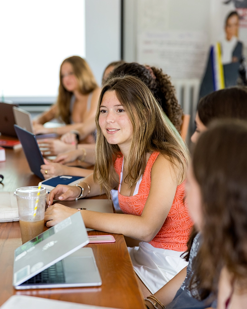 A group of young people sits at a table with laptops and drinks, engaged in discussion. One girl in an orange top is speaking, while others listen attentively.