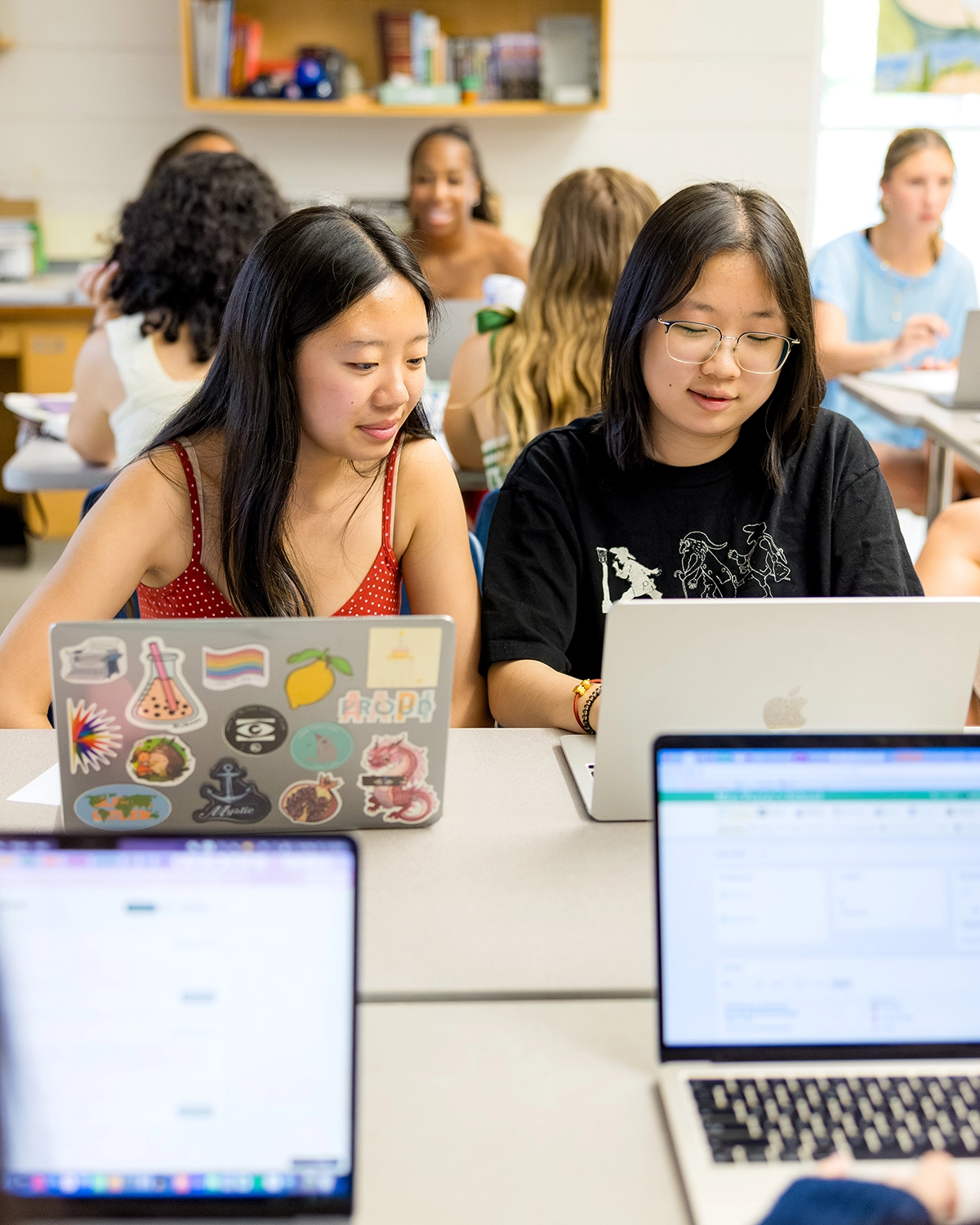 Two students work side by side on laptops at a classroom table, surrounded by classmates, with bookshelves and windows in the background.