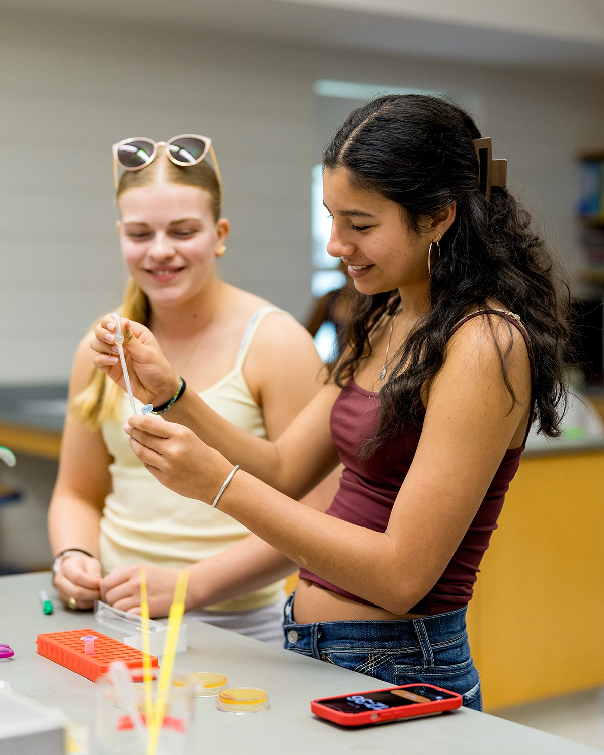 Two young women smile as they work together on a science experiment in a classroom, using pipettes and petri dishes on a lab table.