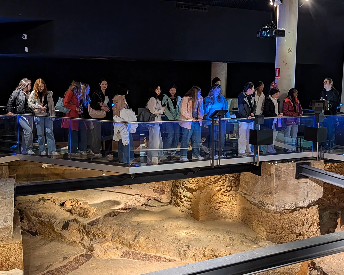 A group of people stand on a glass walkway overlooking ancient ruins in a dimly lit museum or archaeological site.