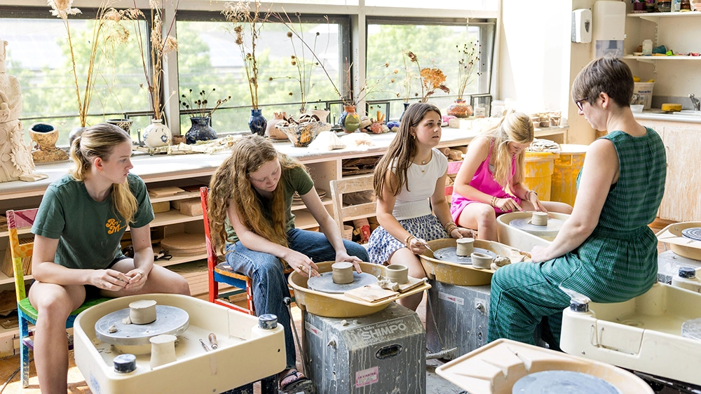 Four students sit at pottery wheels in a brightly lit classroom, shaping clay, while an instructor in a green dress gives guidance. Shelves with art supplies and pottery are visible in the background.