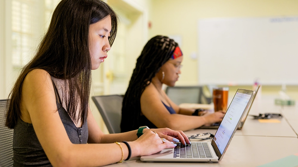 Two women sit at a table working on laptops in a brightly lit room, focusing on their screens. The background includes a whiteboard and windows letting in natural light.