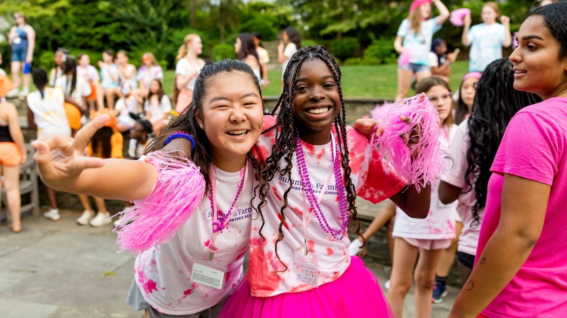 Two girls smiling and posing with pink pom-poms at an outdoor event, surrounded by other kids in colorful outfits, suggesting a fun, festive atmosphere.