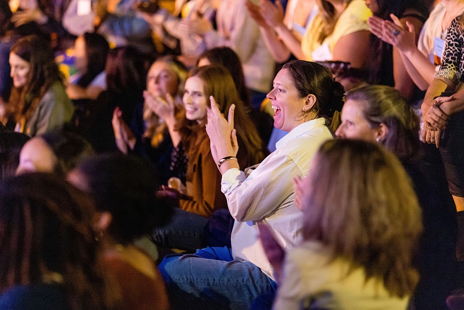 A group of people sit closely together in an audience, clapping and smiling while watching an event or performance. The atmosphere appears lively and engaging.
