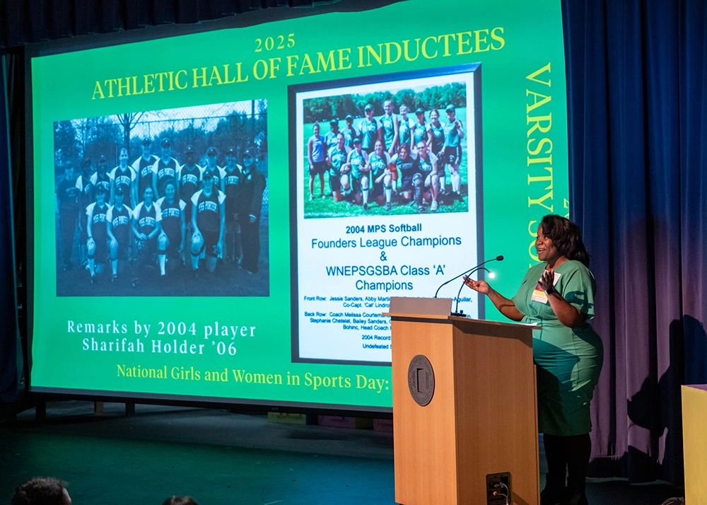 A woman speaks at a podium on stage with a green screen behind her displaying 2025 Athletic Hall of Fame Inductees and images of a softball team.