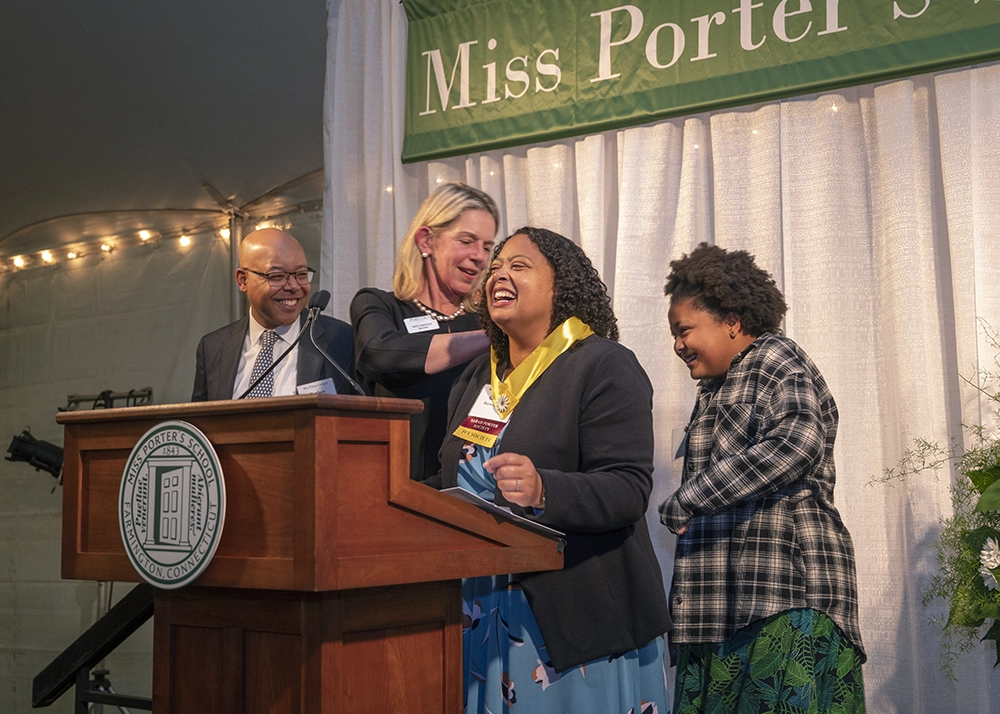 Four people stand at a podium, smiling and laughing. One woman is placing a medal on another woman. A Miss Porters School banner hangs in the background. There are flowers to the right and a seal on the podium.