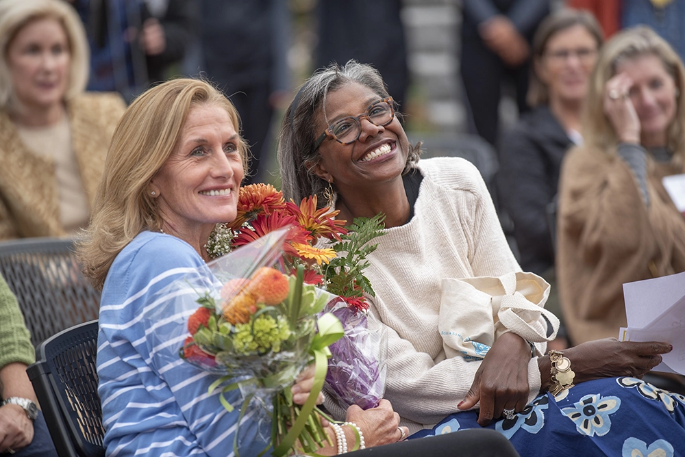 Two women sitting close together, smiling and holding bouquets of colorful flowers, surrounded by other people at an outdoor event.