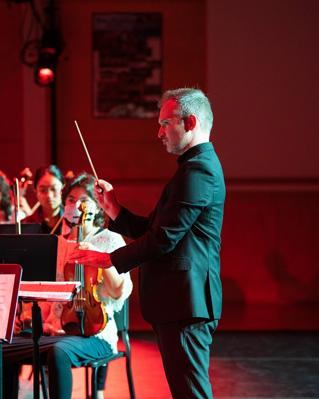 A conductor in a black suit leads an orchestra, holding a baton, while musicians with violins sit and watch attentively on stage under red lighting.