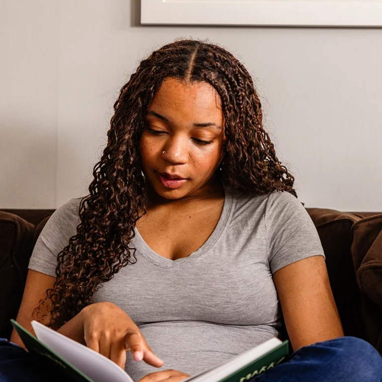 A woman with long curly hair sits on a brown couch, wearing a gray t-shirt, and reads a book, pointing to a page with her finger.
