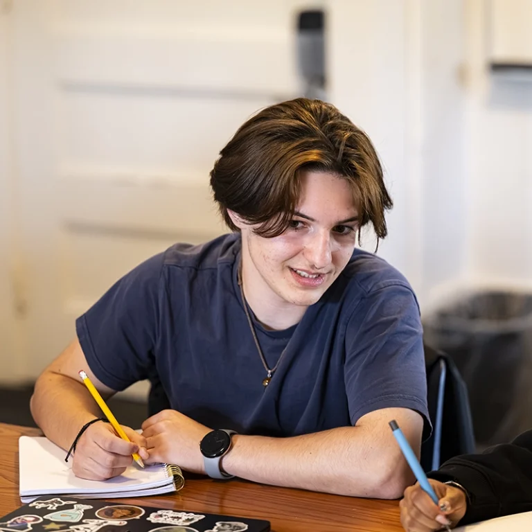 A young person with brown hair and a blue shirt sits at a table, smiling while writing in a notebook. A laptop with stickers is visible in front of them, with another persons hand holding a pen in the foreground.