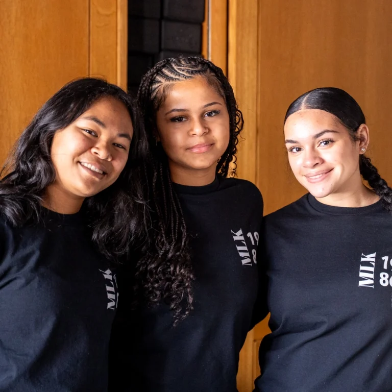Three young women smile, standing close together indoors, wearing matching black shirts with MLK 1988 written on them. A wooden background is behind them.