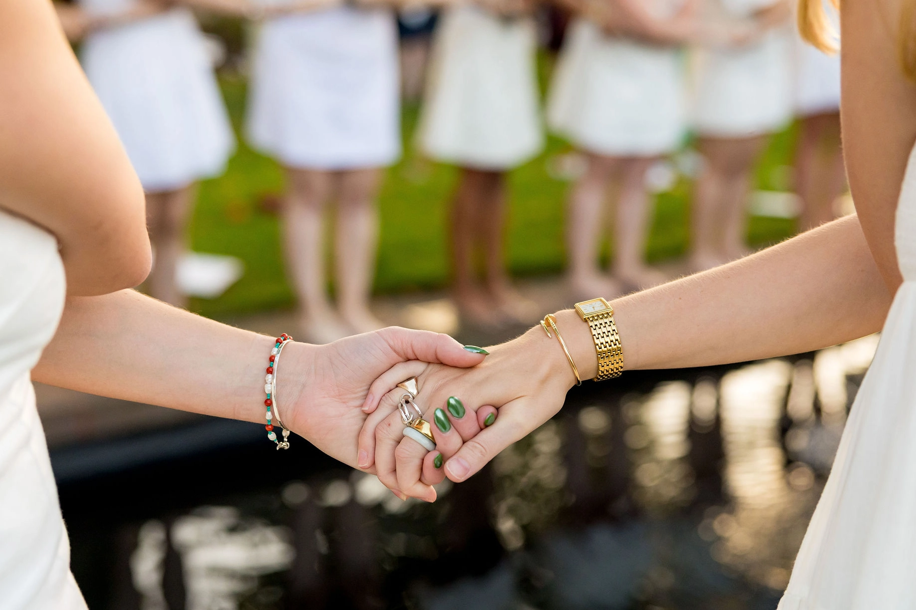 Two people holding hands closely, both wearing white outfits. One has a gold watch and green nail polish; the other has a beaded bracelet and rings. A group in white dresses stands blurred in the background.