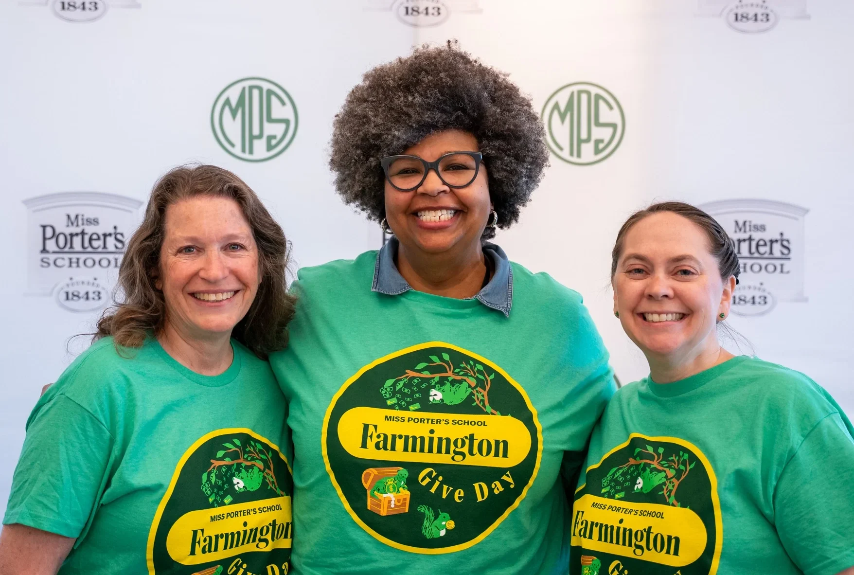 Three smiling women wearing matching green Farmington Give Day t-shirts stand together in front of a Miss Porters School backdrop.