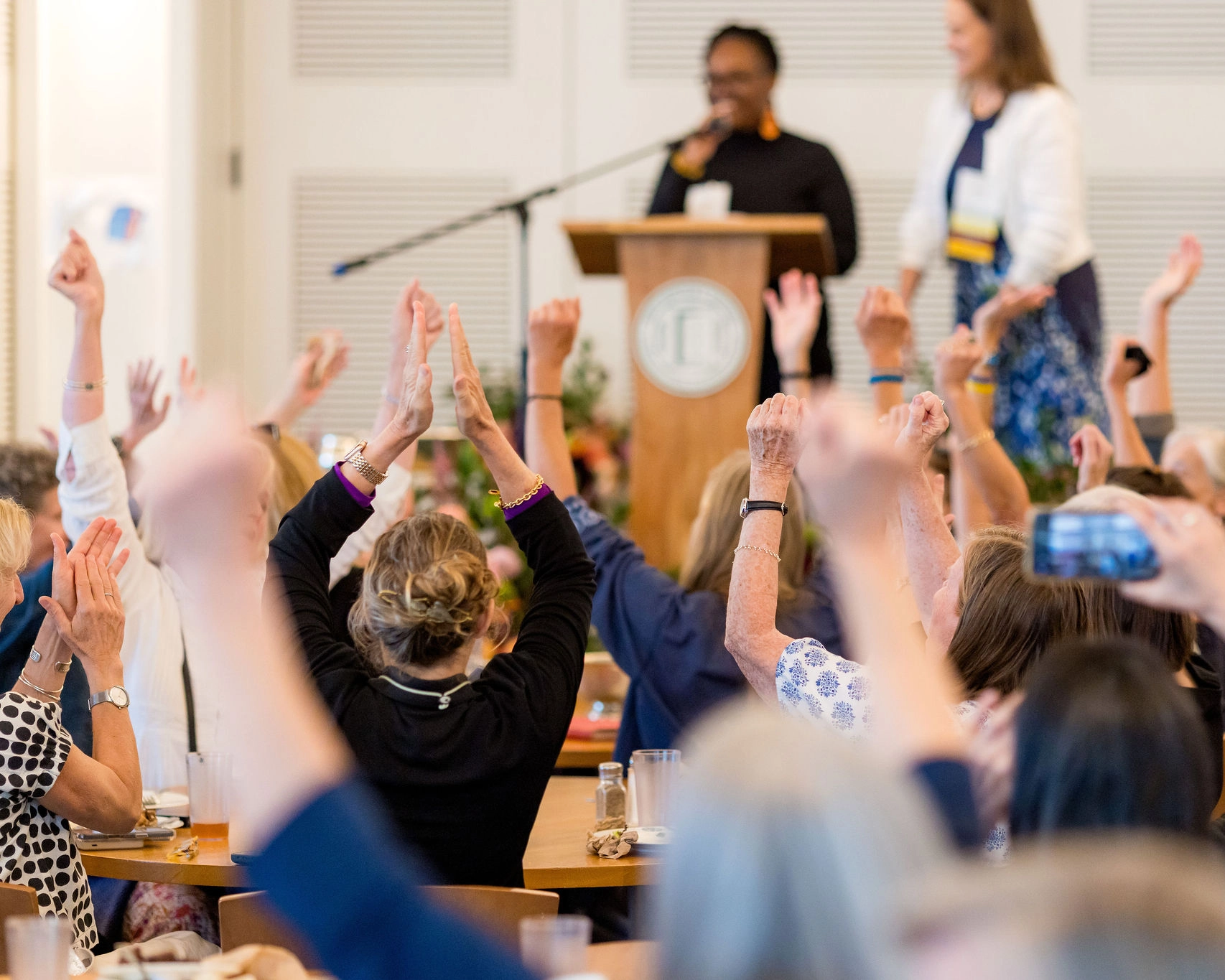 A group of people seated at tables raise their hands in the air, facing a podium where two women stand, one speaking into a microphone. The scene suggests an enthusiastic or celebratory event.