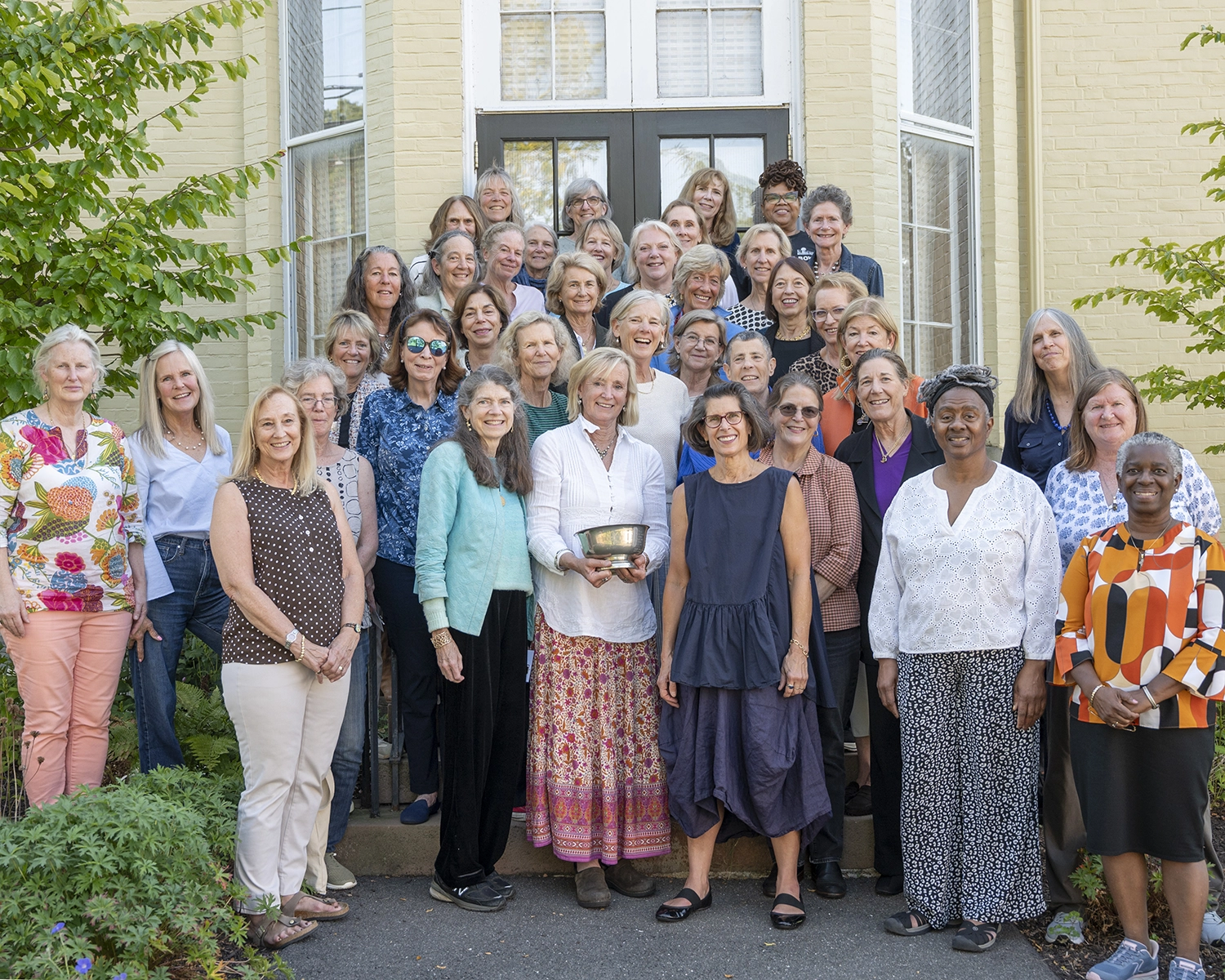 A large group of women of various ages poses together, smiling, in front of a brick building. One woman in the front center holds a silver bowl. Green plants frame the group on either side.