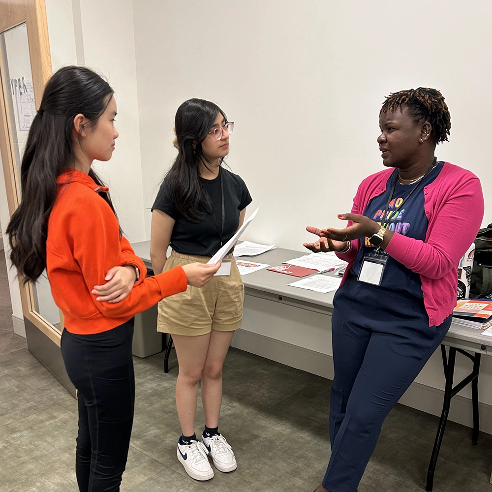 Three women stand talking indoors near a table with papers. One woman in pink gestures while the other two, holding papers, listen attentively. They appear to be having a discussion or interview.