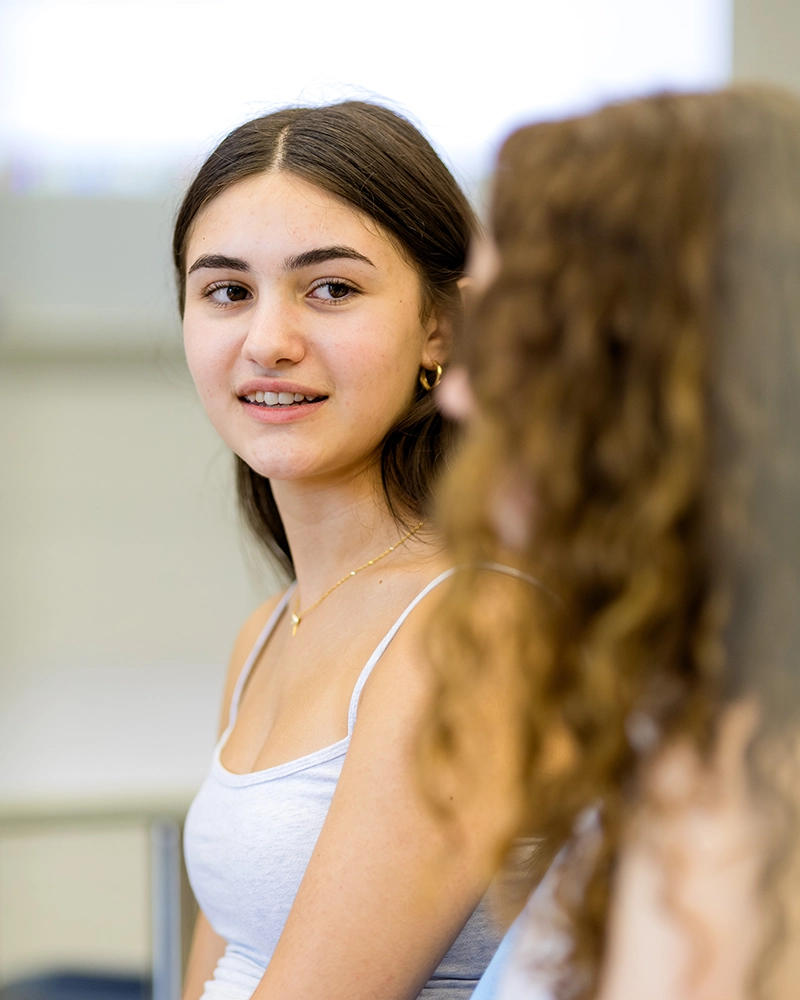 A young woman with long brown hair and a white tank top looks at another person with curly hair, both sitting indoors in a bright room.