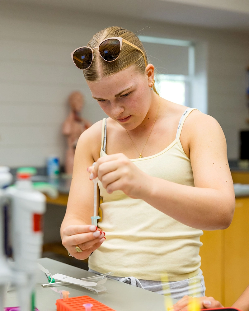 A young person in a yellow tank top and sunglasses on their head uses a pipette in a science lab, focusing on transferring liquid into a small container.