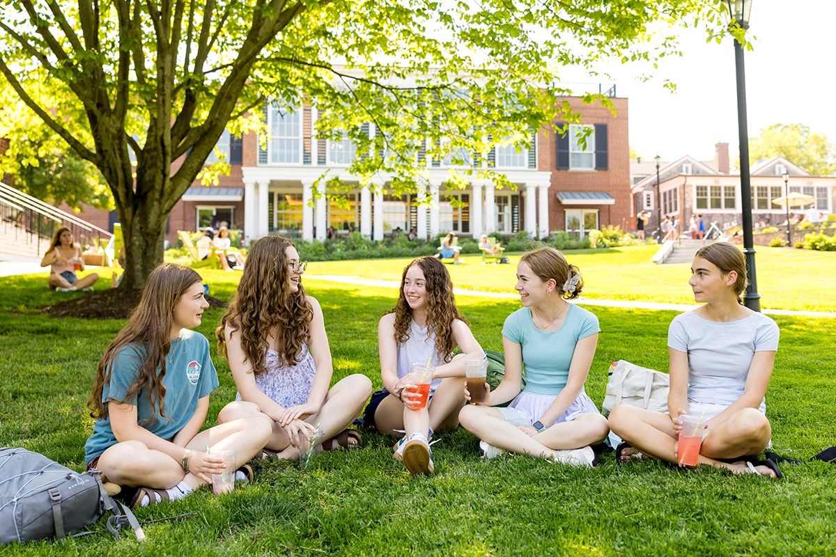 Five young women sit on grass in a circle, talking and holding drinks, with a large tree and a red-brick campus building in the background on a sunny day.