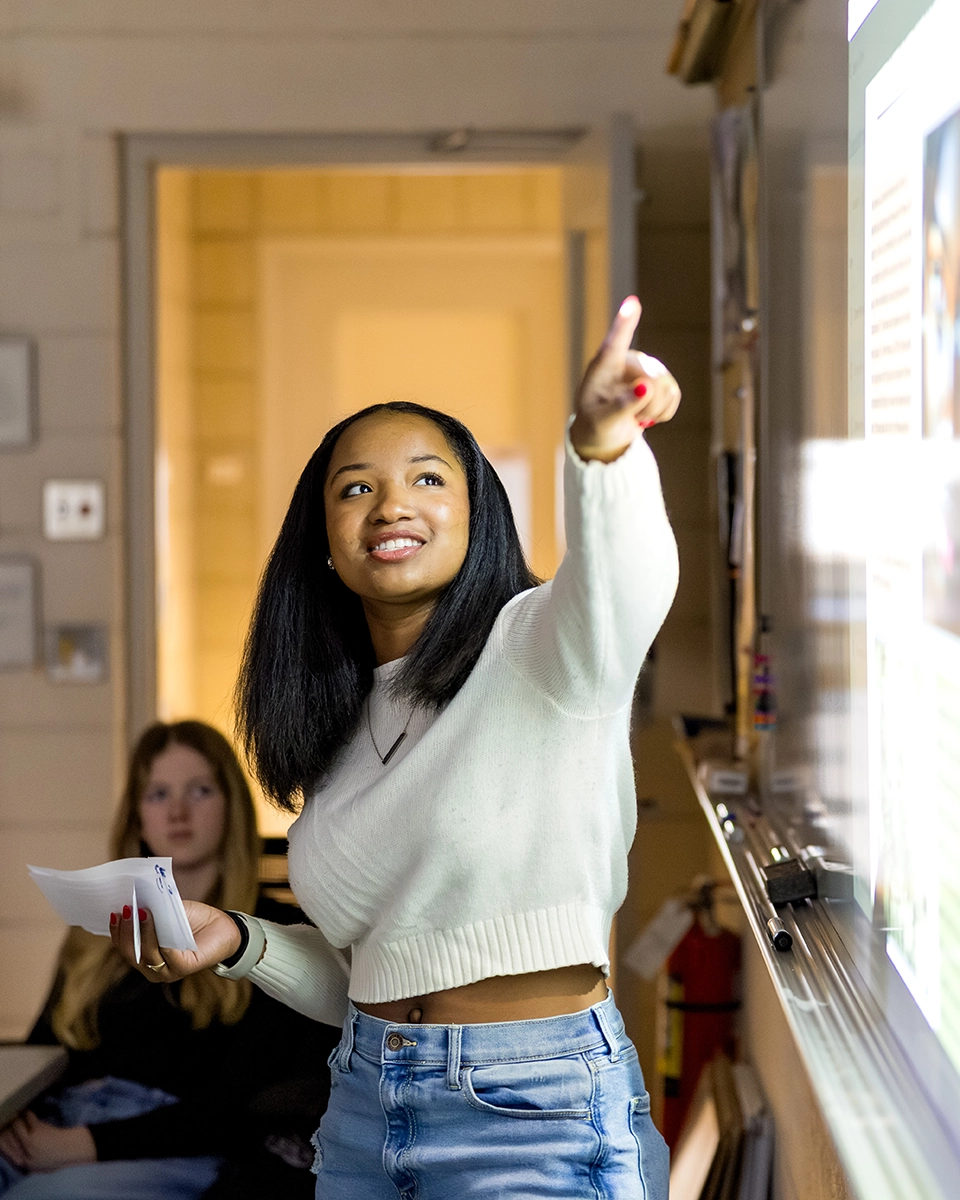 A young woman in a white sweater and jeans smiles while pointing at a projection screen, holding papers in her other hand. Another student sits in the background, watching her presentation.