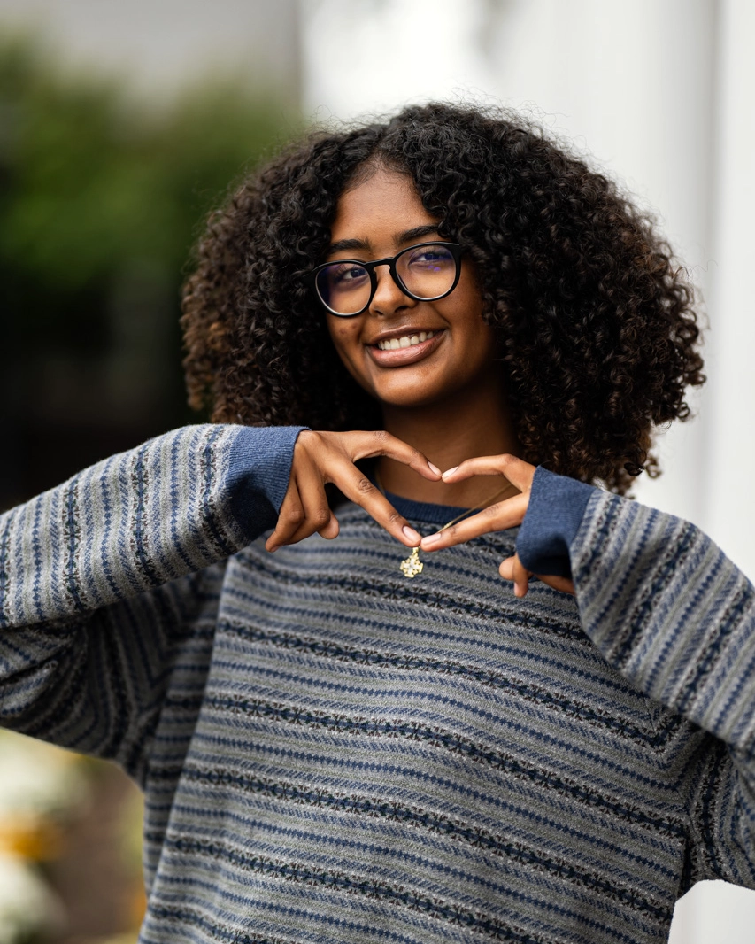 Young person with curly hair and glasses makes a heart shape with their hands while smiling. They wear a striped sweater and stand outdoors, with greenery and blurred flowers in the background.