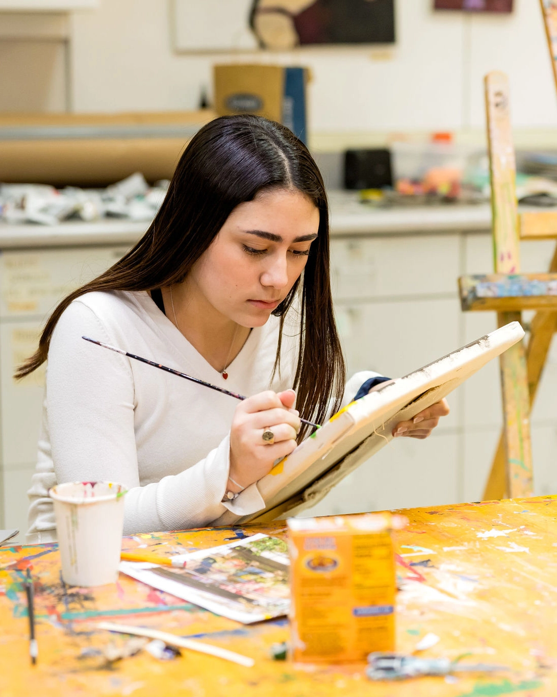 A young woman with long brown hair paints on a canvas at a messy art table, surrounded by art supplies, including brushes, paint, and a drink cup in a brightly lit studio.