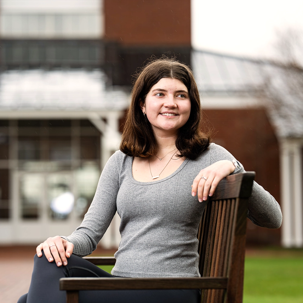 A young woman with shoulder-length brown hair in a gray long-sleeve top sits on a wooden bench outdoors, smiling, with a building and green grass in the background.