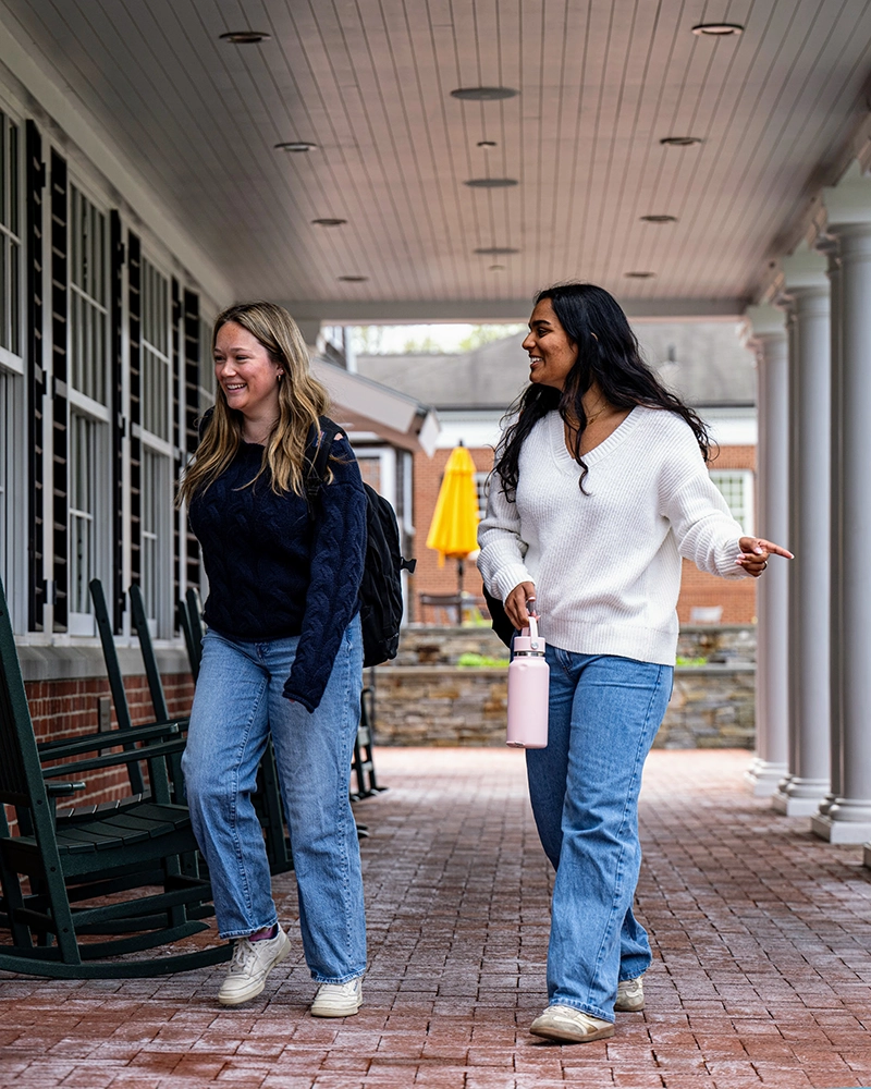 Two young women walk and smile together under a covered walkway with columns. One carries a backpack, and the other holds a pink water bottle. Both wear jeans and sweaters. Outdoor seating and a yellow umbrella are visible behind them.