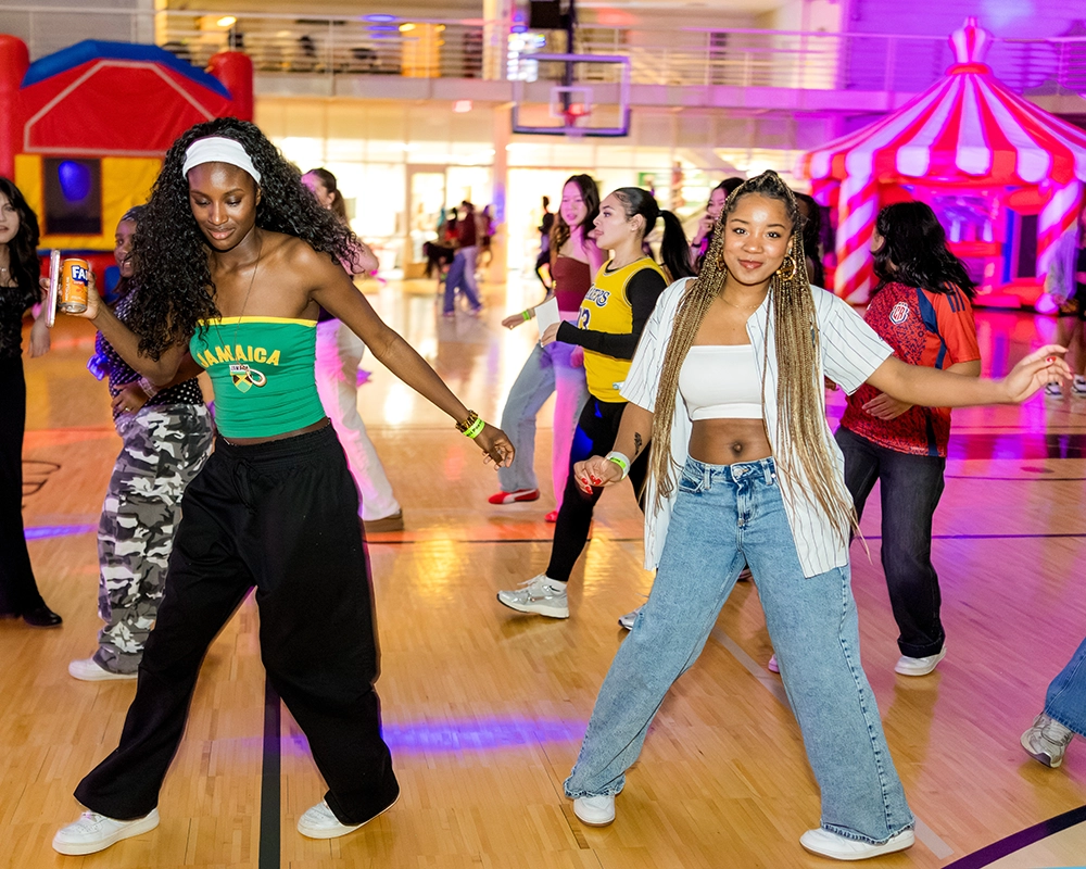 Two young women dance in a gymnasium with others in the background. One wears a green Jamaica tank top, the other a striped shirt and jeans. Colorful lights and inflatables are visible.