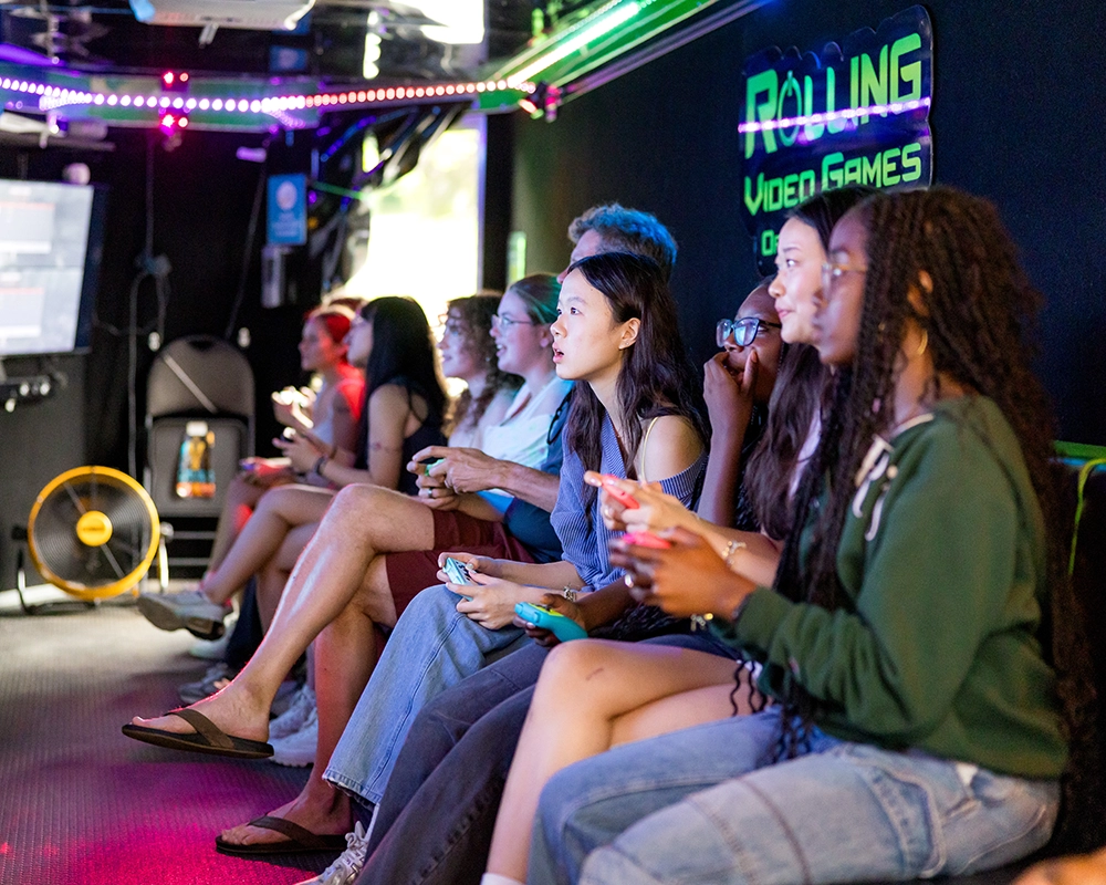 A group of young people sit in a row inside a neon-lit gaming van, focused on playing video games with controllers in their hands.