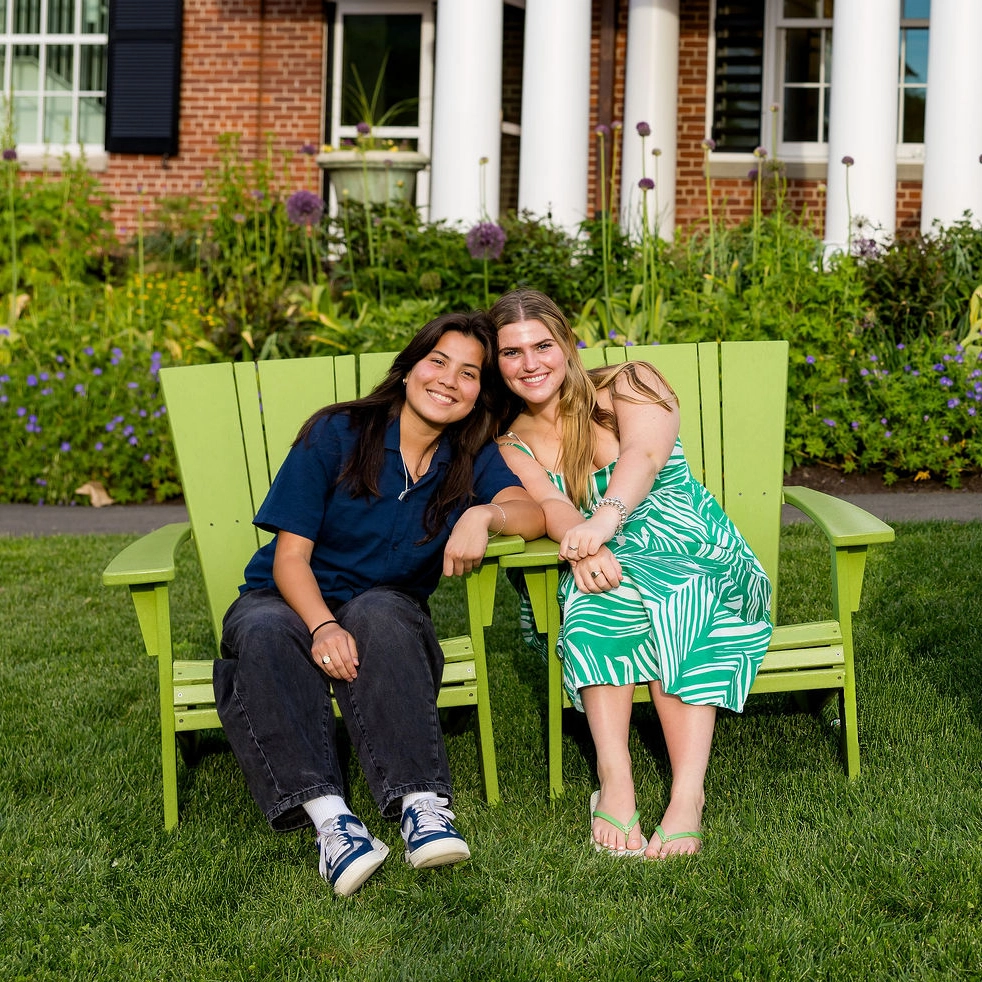 Two people sit closely together on green chairs outside, smiling at the camera. There is a garden with flowers and a building with white columns in the background.