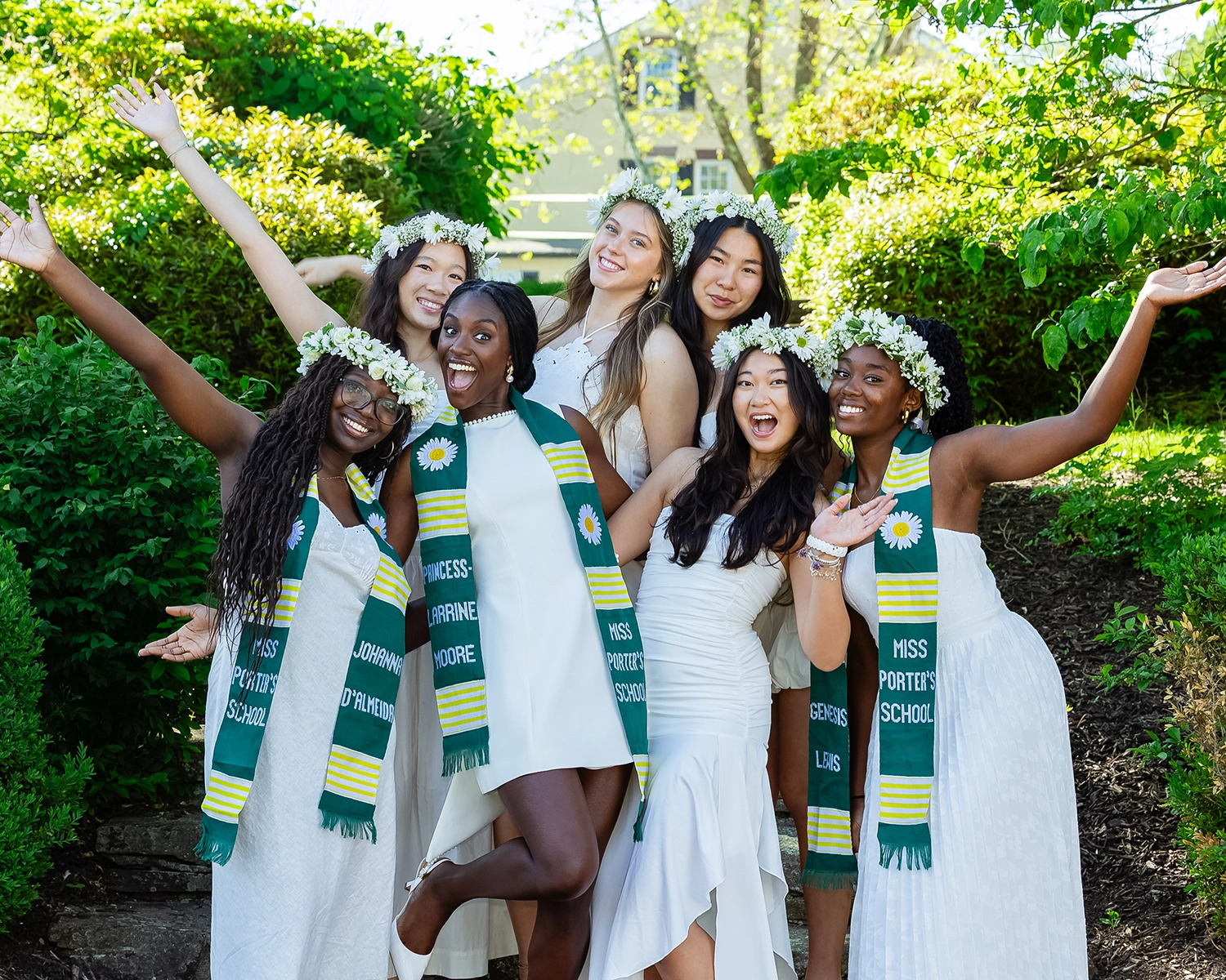 Six young women in white dresses and flower crowns stand outdoors, smiling with arms raised. They wear green and yellow sashes that read Miss Porters School. Lush greenery and a house are visible in the background.