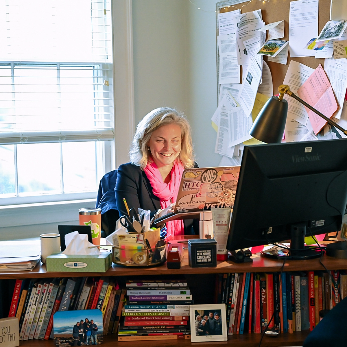 A woman with gray hair and a pink scarf smiles while working at a desk cluttered with books, papers, and a computer, near a sunlit window and a corkboard with pinned documents.
