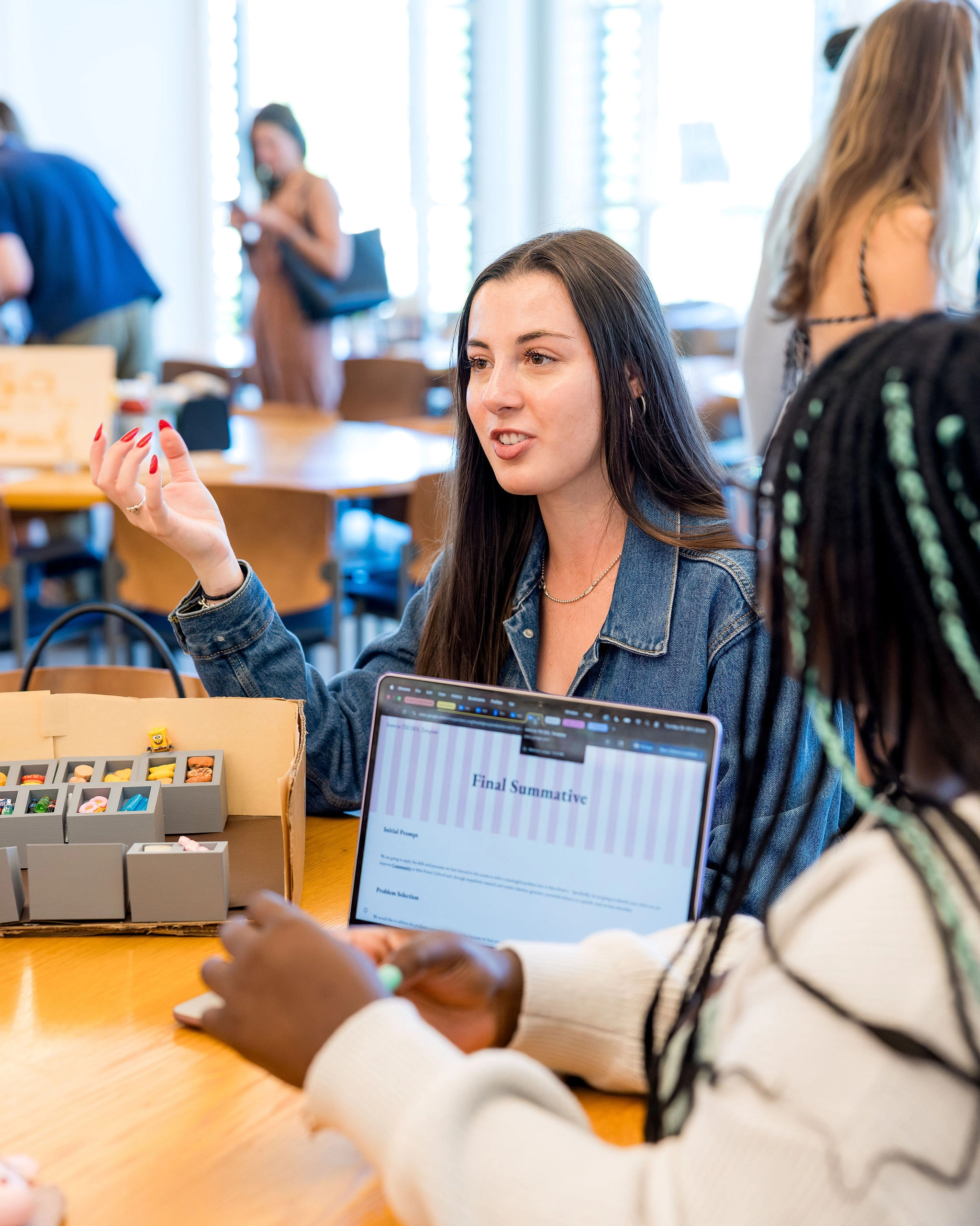 A woman talks at a table with others. In front of her is a laptop displaying Final Summative. Small boxes and papers are on the table, and the setting appears to be a classroom or group workspace.