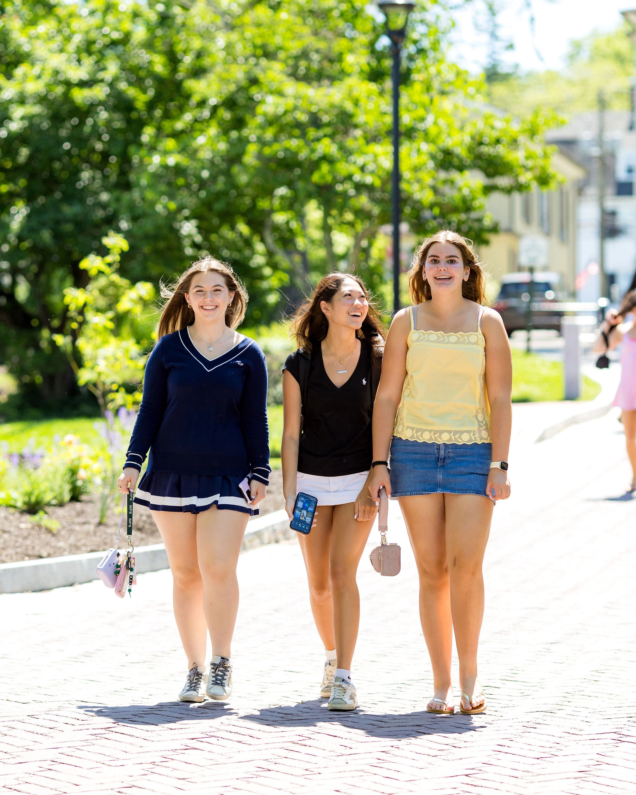 Three young women walk outside on a sunny day, smiling. They wear casual summer outfits and hold small bags or phones, with greenery and buildings in the background.