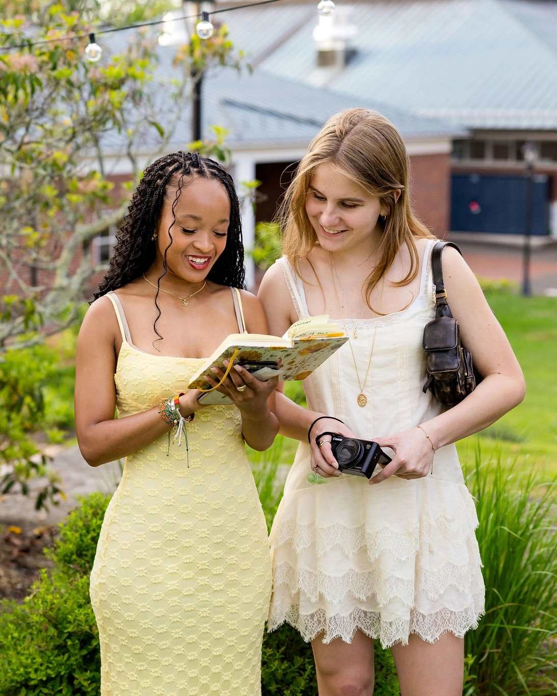 Two women in light dresses stand outside, smiling and looking at an open book. One holds a camera. There is greenery and a building in the background.