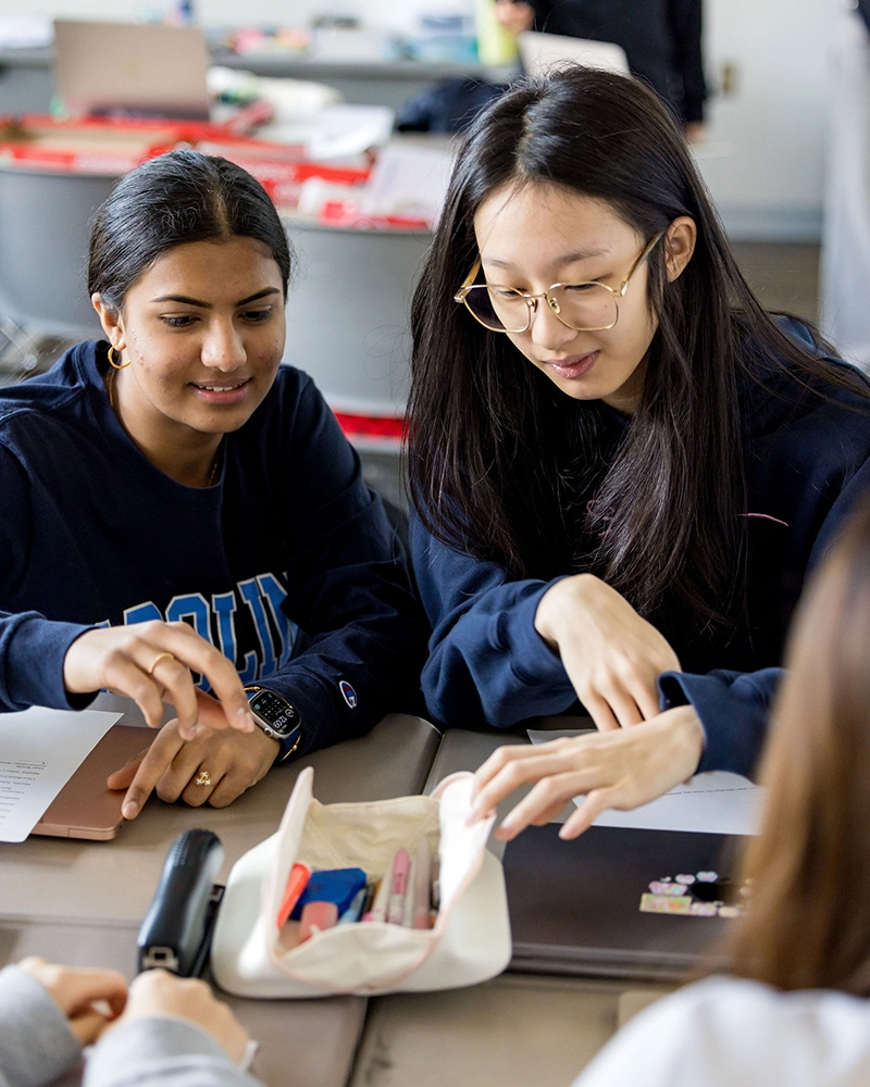 Two students sit at a desk, looking at a pencil case with school supplies. One is pointing at the items inside, while the other smiles. Papers, a laptop, and other items are scattered on the table.