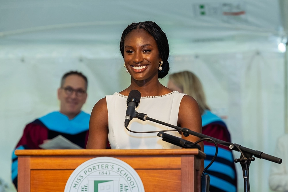A young woman in a white dress smiles while speaking at a podium during a graduation ceremony. People in academic regalia stand behind her. The podium features the Miss Porters School emblem.
