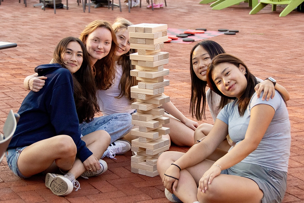 Five young women sit on brick pavement around a tall Jenga tower, smiling at the camera. There are chairs and outdoor games in the background.