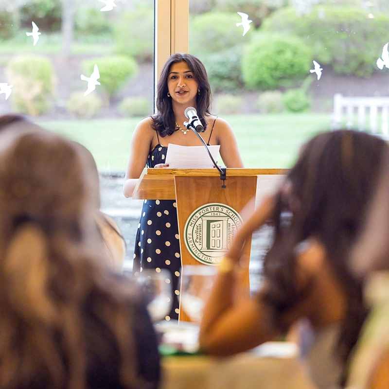 A woman in a black polka dot dress speaks at a podium with a seal, addressing an audience in a bright room with large windows and bird decorations.