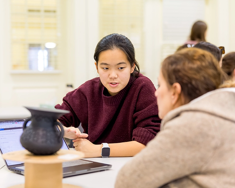 Two people sit at a table, one gesturing toward a laptop while talking. A dark vase sits between them. They appear to be discussing something in a bright indoor setting.