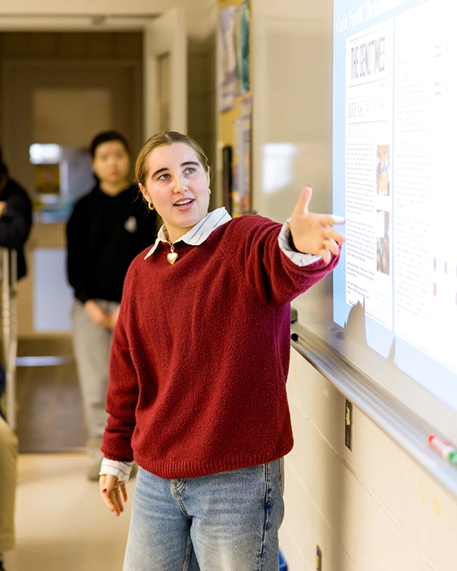 A person in a red sweater gestures toward a projected presentation on a classroom whiteboard, speaking to others while students watch in the background.