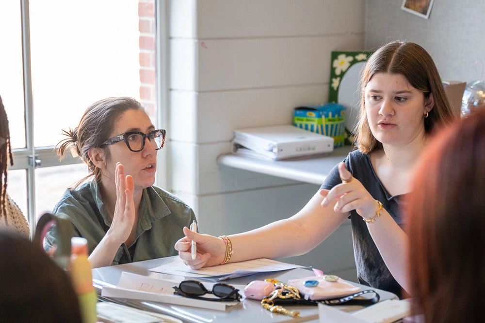 Two women sit at a table with papers and items, talking and gesturing. One raises her hand, the other points. Sunlight comes through a window behind them.