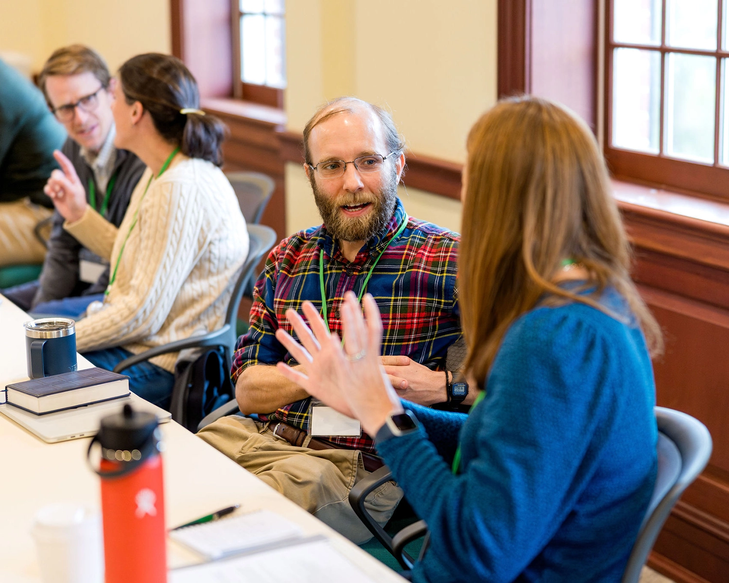 Four people sit at a table in a bright room, engaged in conversation. The man in the center with glasses and a plaid shirt talks animatedly with a woman facing him. Books, water bottles, and papers are visible on the table.