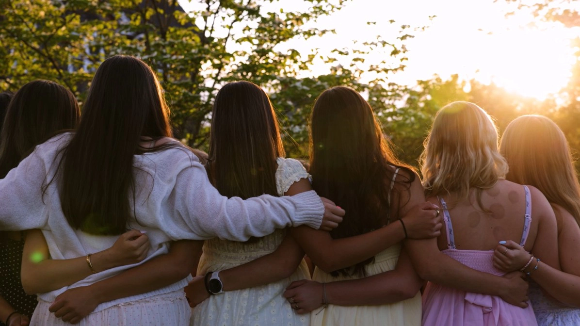 Six Porter's girls stand outdoors with their backs to the camera, arms around each other, in dresses and casual clothes, as the sun sets behind them, creating a warm and supportive atmosphere.