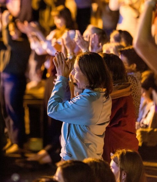 A woman in a light jacket stands among a crowd, clapping enthusiastically with both hands raised, while others around her also applaud. The group is bathed in warm, golden light.