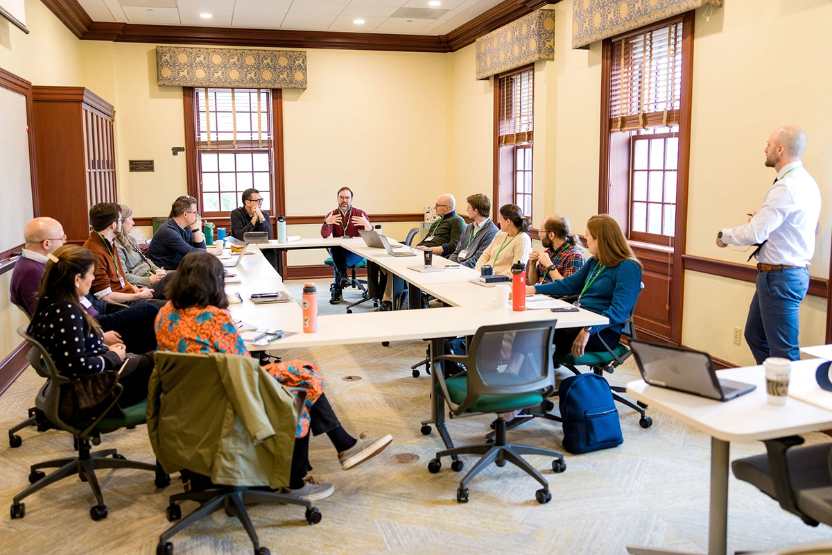 A group of people sit around a U-shaped table in a conference room, listening to a man who is standing and speaking about opportunities for educators. Laptops, notebooks, and drinks are on the tables.