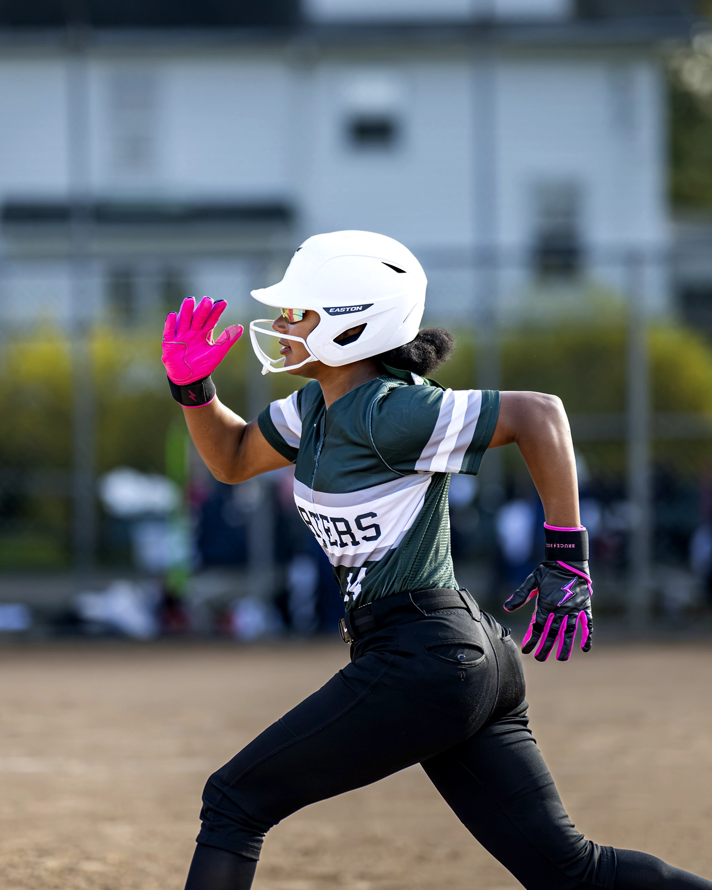 A softball player in a helmet and pink gloves sprints on a dirt field, with a fence and blurred buildings in the background.
